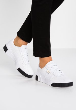 Sneakers bianche Puma con suola nera indossate con pantaloni neri su un pavimento grigio chiaro.