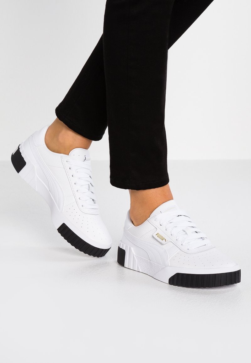 Baskets blanches Puma avec semelles noires portées avec un pantalon noir sur un sol gris clair.