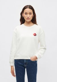 Felpa color crema chiaro in misto cotone; presenta una grafica di labbra rosse sul petto. Il design include una scollatura rotonda e polsini a costine.