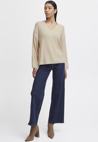 Beige bordás kötött pulóver v-nyakkivágással, sötétkék, bő szárú farmernadrággal párosítva. A modellen bézs, hegyes orrú bokacipő van.