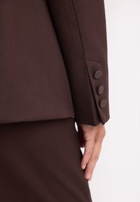 Brąsowy garnitur o gładkiej fakturze, z czterema ciemnymi guzikami na rękawie i dopasowanym krojem. Zbliżenie na rękaw i dłoń.