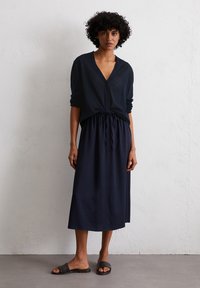 Cardigan bleu marine à boutons superposé sur une jupe bleu marine longueur cheville, nouée à la taille ; le mannequin porte des sandales à glisser foncées. Tissu lisse, coupe décontractée.