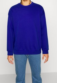 Persona che indossa una felpa semplice blu reale con maniche lunghe e jeans di denim blu chiaro, in piedi davanti a uno sfondo chiaro e semplice.