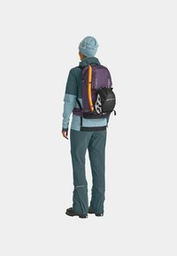 Back view of a person wearing a blue-green outfit with a purple backpack. Backpack features an orange tool attached, with black accents.