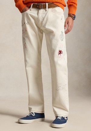 Άνδρας με παντελόνι κρεμ με κεντημένα σχέδια, μπλε αθλητικά παπούτσια, καφέ ζώνη, πορτοκαλί πουλόβερ και ρολόι στον αριστερό καρπό.