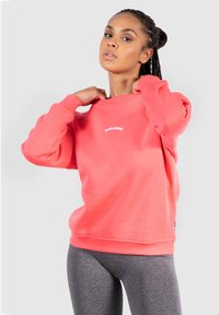 Korallfärgad tröja med ribbade ärmslut och fåll, med en vit logga på framsidan. Matchad med grå leggings. Slät textur, avslappnad design.