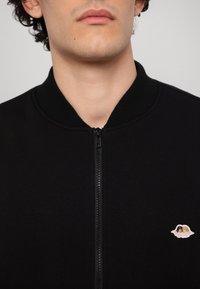 Juodas zip-up bomber tipo švarkas su įmegztu apykakle. Pastebimas siuvinėtas logotipo detalė dešiniajame apatiniame kampe. Lygi audinio tekstūra.