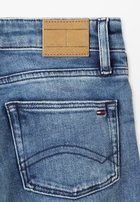 Vaqueros de mezclilla azul claro con una etiqueta de cuero marrón en la cintura, detalles con costuras y un bolsillo trasero que presenta un pequeño logo en rojo, blanco y azul.