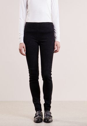 Osoba ubrana w czarne dopasowane spodnie, białą bluzkę z długim rękawem oraz czarne buty z klamrami i ćwiekami, stojąca na jednolitym tle.