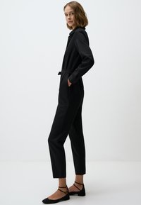 Zwarte jumpsuit met een riem in de taille, lange mouwen en een kraag. Heeft zijzakken en taps toelopende benen. Gemaakt om te dragen met zwarte sandalen met bandjes.