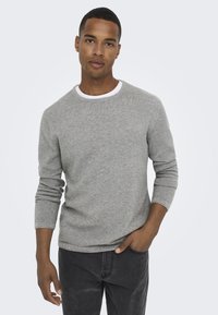 Maglione grigio a maglia con scollo rotondo e maniche lunghe, con un motivo testurizzato e un'accenno di colletto bianco, abbinato a jeans scuri.