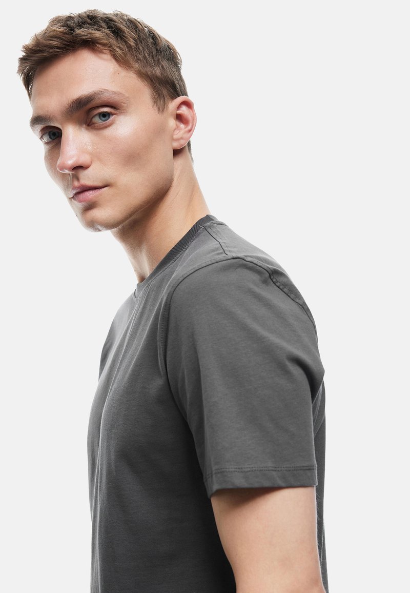 Mørkegrå t-shirt med rund hals, der har korte ærmer, blødt bomuldsstof og synlige syninger langs sømmene. Modellen vises i profil.