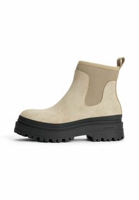 Beige ruskind ankelstøvler med teksturerede elastiske sidelapper og sort gummisål med dybe trækkrofler. Håndtag på bagsiden.