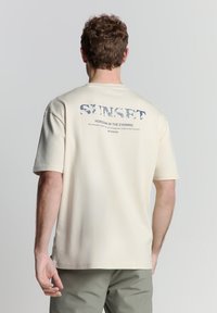 Beige katoenen T-shirt met ronde hals, korte mouwen en een grafische print op de achterkant met tekst in blauw en grijs.