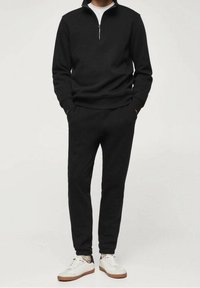 Maglione nero con collo a zip e bordo a coste, abbinato a pantaloni affusolati coordinati. Completa il look un paio di sneakers bianche con suola marrone chiaro.