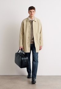 Μπεζ, υπερμεγέθη αδιάβροχο με κούμπωμα από κουμπιά, φορεμένο πάνω από ένα ανοιχτό πουλόβερ και σκούρο τζιν. Το μοντέλο κρατάει μια μαύρη δερμάτινη τσάντα ταξιδιού.