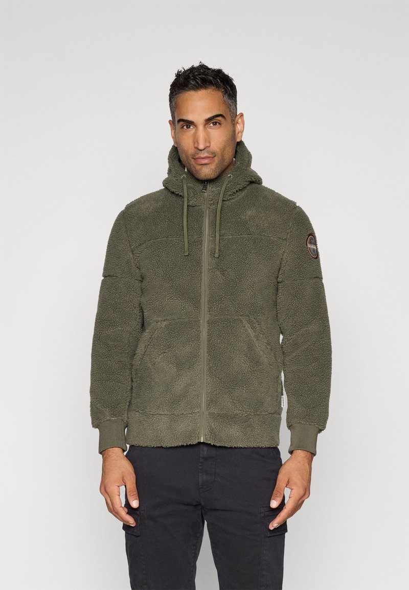 Mann trägt eine olivgrüne Fleecejacke mit Reißverschluss und Kapuze, mit Kordelzügen, und steht vor einem schlichten hellen Hintergrund.