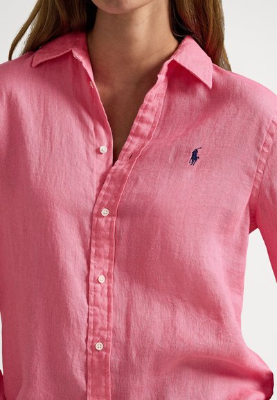 Sieviete tērpusies rozā pogājām aizpogājamā linu kreklā ar apkakli un tumši zilu izšūtu polo spēlētāja logotipu uz krūtīm.