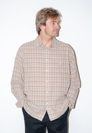 Mand iført beige og hvid ternet langærmet skjorte med hænderne i lommerne, kigger mod højre mod en ensartet hvid baggrund.