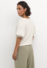 Cremefarbenes Strickoberteil mit Puffärmeln, rundem Ausschnitt, subtiler Textur, kombiniert mit hellgrünen hochgeschnittenen Hosen und einem goldenen Armband.
