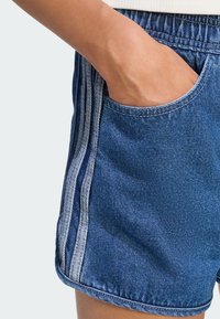 Ręka spoczywa wewnątrz kieszeni niebieskich szortów dżinsowych z dwoma pionowymi białymi paskami po bokach.