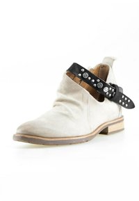 Bottine beige clair en daim avec talon bas en bois et bride en cuir noir ornée de clous en argent et de décorations circulaires.