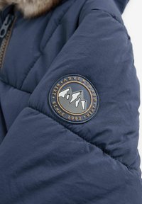 Granatowa kurtka puchowa z okrągłą, wytłoczoną naszywką z górskim motywem i napisem "ADVENTURER SINCE 1982."