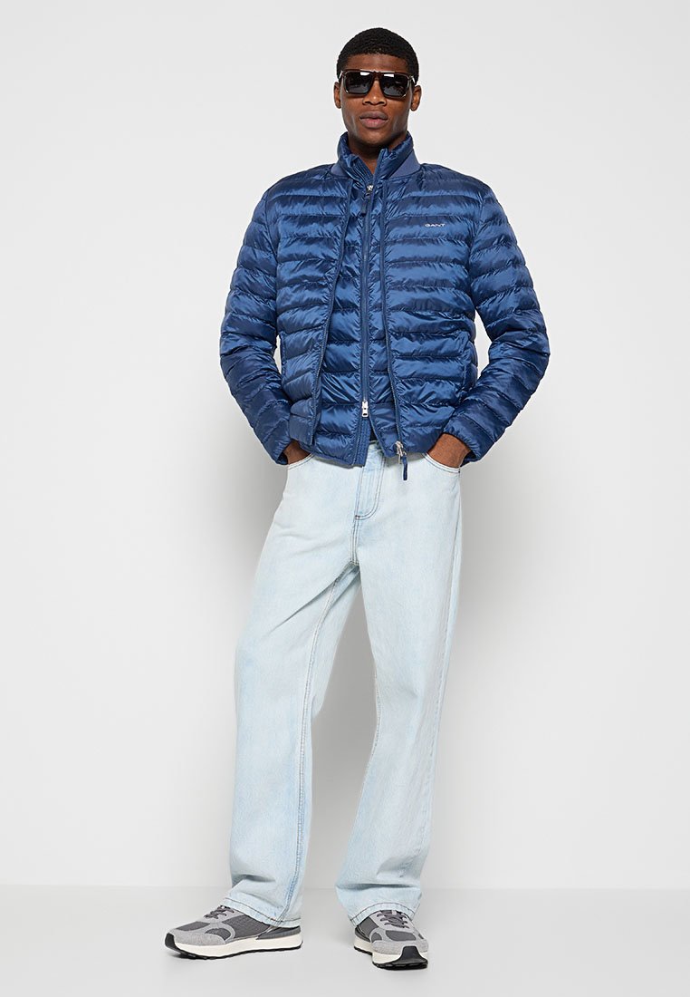 Granatowa ocieplana kurtka z zamkiem błyskawicznym i podniesionym kołnierzem, zestawiona z jasnoniebieskimi dżinsami i szarymi trampkami. W okularach przeciwsłonecznych.