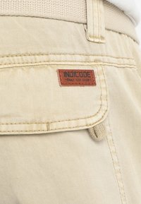 Gros plan d'une poche en tissu beige avec une étiquette marron portant la mention "INDICODE Wear the code" et un bouton recouvert de tissu sous le rabat de la poche.