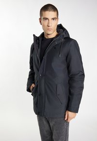 Giacca nera con cappuccio dal design liscio, con tasche zip, chiusura a bottone e un logo discreto, abbinata a jeans grigi.
