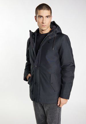 Giacca nera con cappuccio dal design liscio, con tasche zip, chiusura a bottone e un logo discreto, abbinata a jeans grigi.