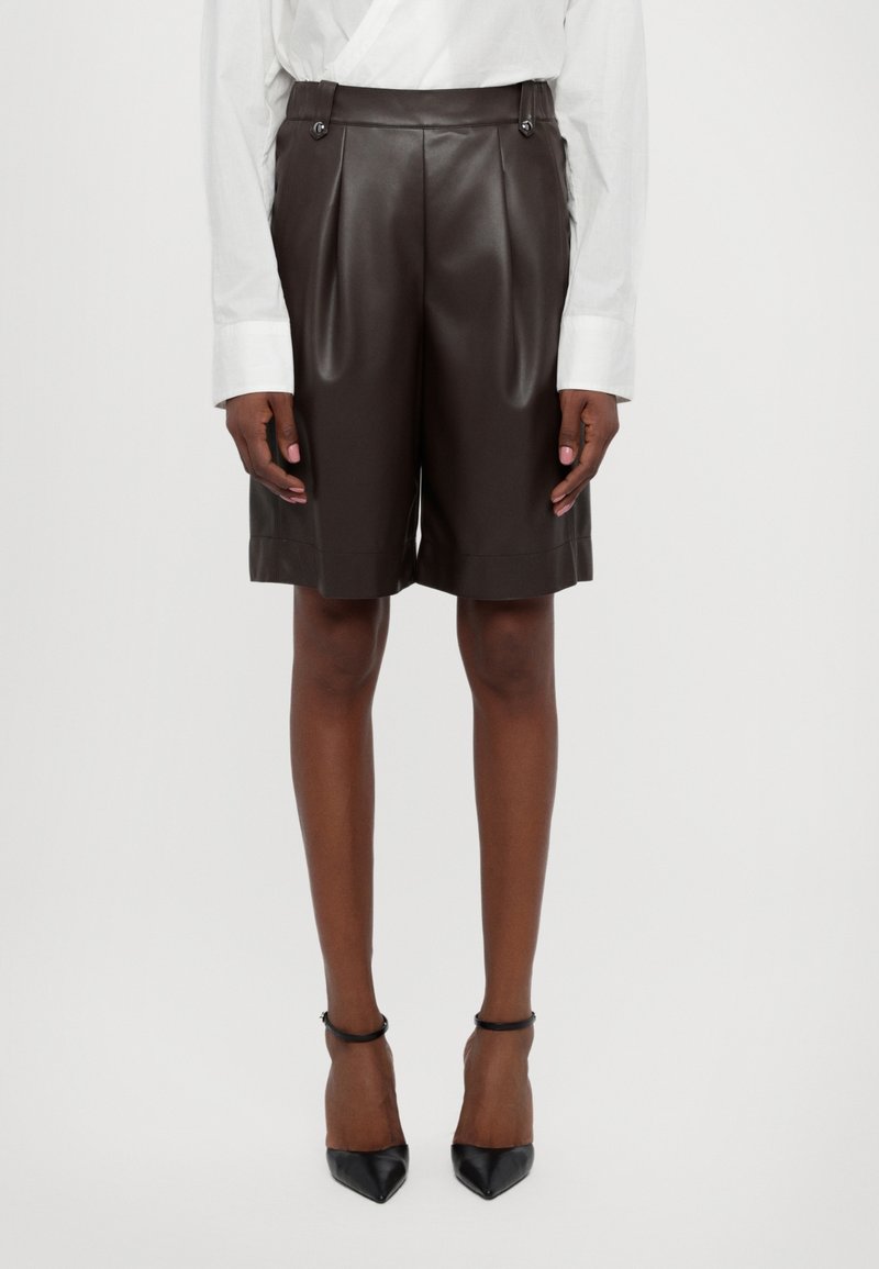 Personne portant un short en cuir marron foncé taille haute, une chemise blanche à manches longues et des talons hauts noirs à bout pointu avec des brides à la cheville.
