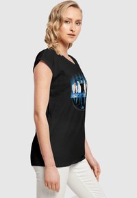 Schwarzes T-Shirt mit kurzen Ärmeln, das ein kreisförmiges Grafikdesign von fünf Personen und den Text "Backstreet" in Blau zeigt, vor einem kontrastierenden Hintergrund.