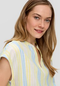 Chemise légère à manches courtes, col montant, ornée de rayures verticales jaunes et bleues sur un fond blanc. Tissu doux et texturé.