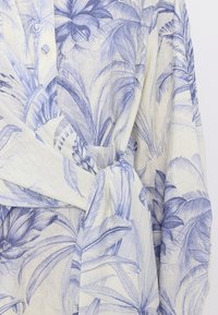 Cămașă ușoară din in, cu un model tropical de frunze de palmier albastre pe un fundal deschis, având o croială lejeră și un detaliu de legare în jurul taliei.