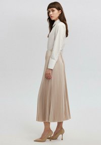 Kermansblouse, jossa on terävä kaulus, yhdistetty polvenpituiseen laskostettuun beigeen hameeseen. Mallilla on päällään beigeet teräväkärkiset korkokengät. Minimalistinen muotoilu.
