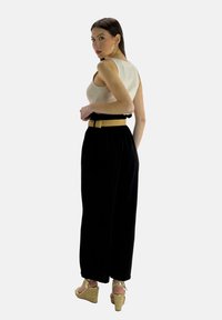 Zwarte wijde broek met een gestructureerde gouden riem, gecombineerd met een aangesloten lichtbeige tanktop. Het model staat zijwaarts met een lichte draai.