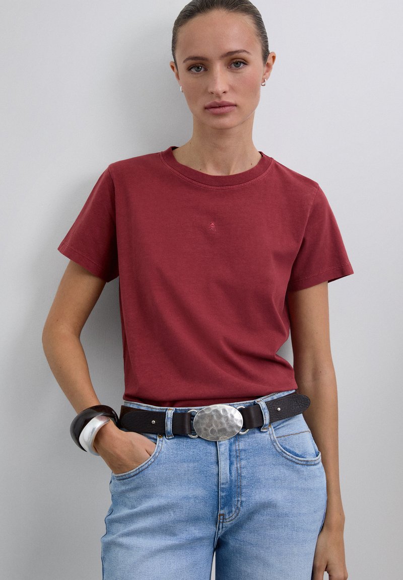 T-shirt en coton rouge avec un col rond classique, associé à un jean bleu clair et une ceinture noire avec une boucle argentée texturée.