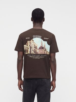 Homme portant un t-shirt marron avec un paysage urbain vintage et un texte au dos, assorti à un jean en denim foncé.
