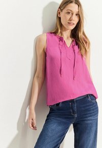 Rosa ärmlös topp med V-ringning och dragsko-detalj, kombinerad med mörkblå jeans. Tyget verkar lätt och har en mjuk textur.