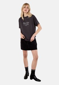 Dunkelgraues T-Shirt mit dem Text "Peace & Love CLUB", kombiniert mit einem schwarzen Jeansrock und schwarzen Chelsea-Stiefeln, stehend vor einem neutralen Hintergrund.