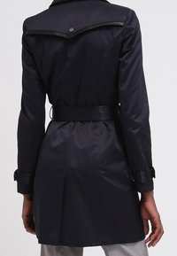 Dark navy trench coat with a belted waist, flap collar, and glossy accents. Made from smooth material with a fitted silhouette and cuff details.