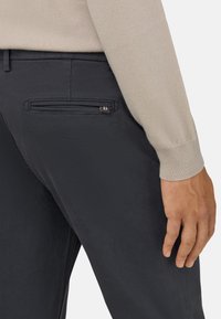 Gaiši pelēki bikses ir izgatavotas no teksturēta auduma, ar kabatu uz aizmugures, bet virs tām redzams gaiši bežs džemperis.