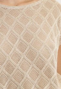 Haut tricot beige clair avec un motif en maille diamant et tissage ouvert. Le tissu a une texture douce et une coupe décontractée.