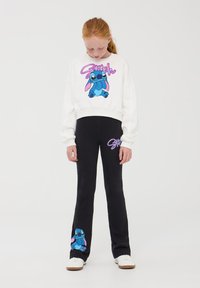 Felpa bianca con un design di personaggio cartoon blu, abbinata a pantaloni neri a zampa d'elefante con una grafica del personaggio abbinata. Sneakers bianche.