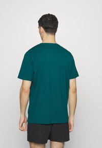 Turkusowa bawełniana koszulka z okrągłym dekoltem, krótkimi rękawami i prostym krojem, widoczna z tyłu. Na dole widać czarne szorty.