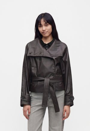 Jeune femme aux longs cheveux foncés portant une veste en cuir marron foncé avec ceinture et un pantalon gris clair sur un fond blanc uni.
