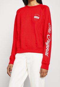 Czerwony sweatshirt z długimi rękawami, z białym graficznym napisem na rękawie i klatce piersiowej. Wykonany z bawełny, o luźnym kroju.
