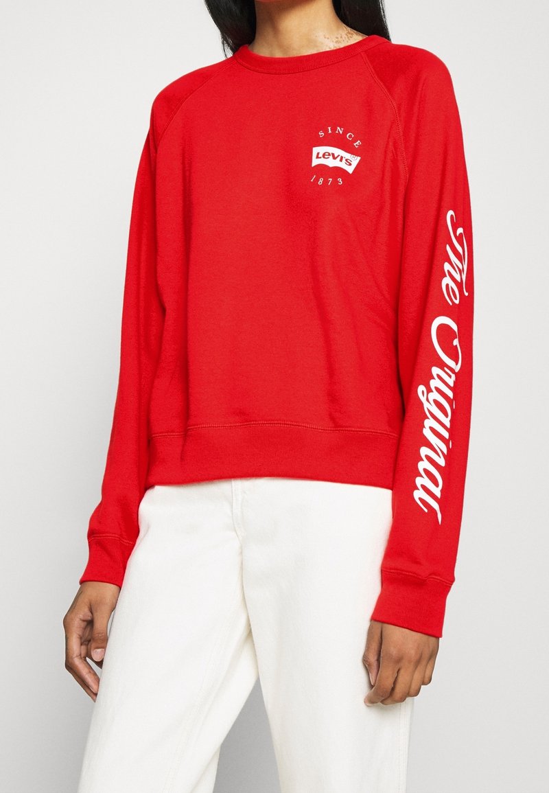 Czerwony sweatshirt z długimi rękawami, z białym graficznym napisem na rękawie i klatce piersiowej. Wykonany z bawełny, o luźnym kroju.