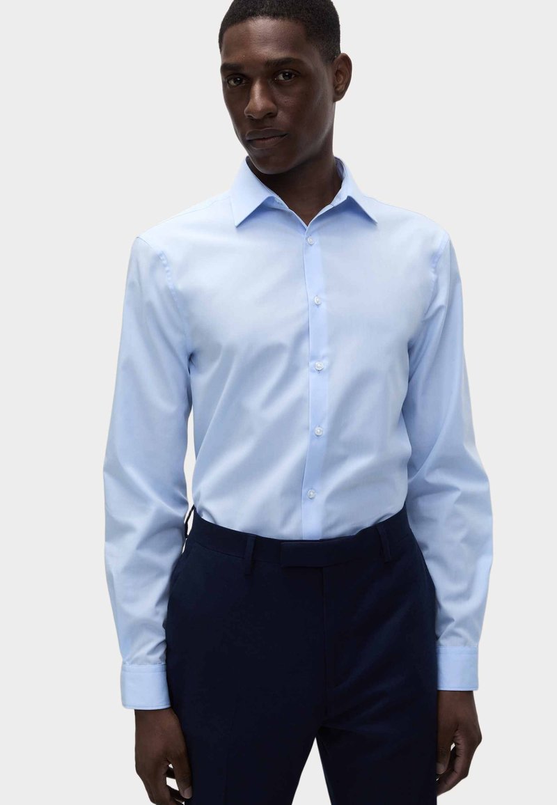 Camicia azzurro chiaro con colletto classico e maniche lunghe, caratterizzata da una texture liscia e una chiusura con bottoni. Abbinata a pantaloni scuri.